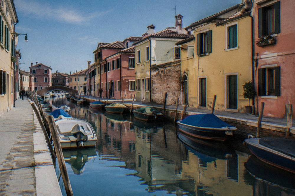 A narrow canal with moored small boats and pastel houses reflected in still water