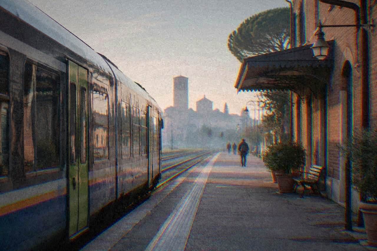 A regional train parked at a platform beside a small Italian railway station building