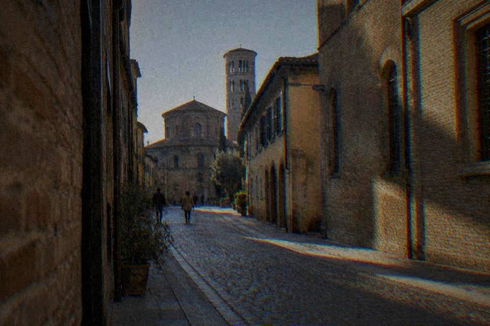 Passengers with suitcases walking from a station forecourt towards a row of Italian townhouses