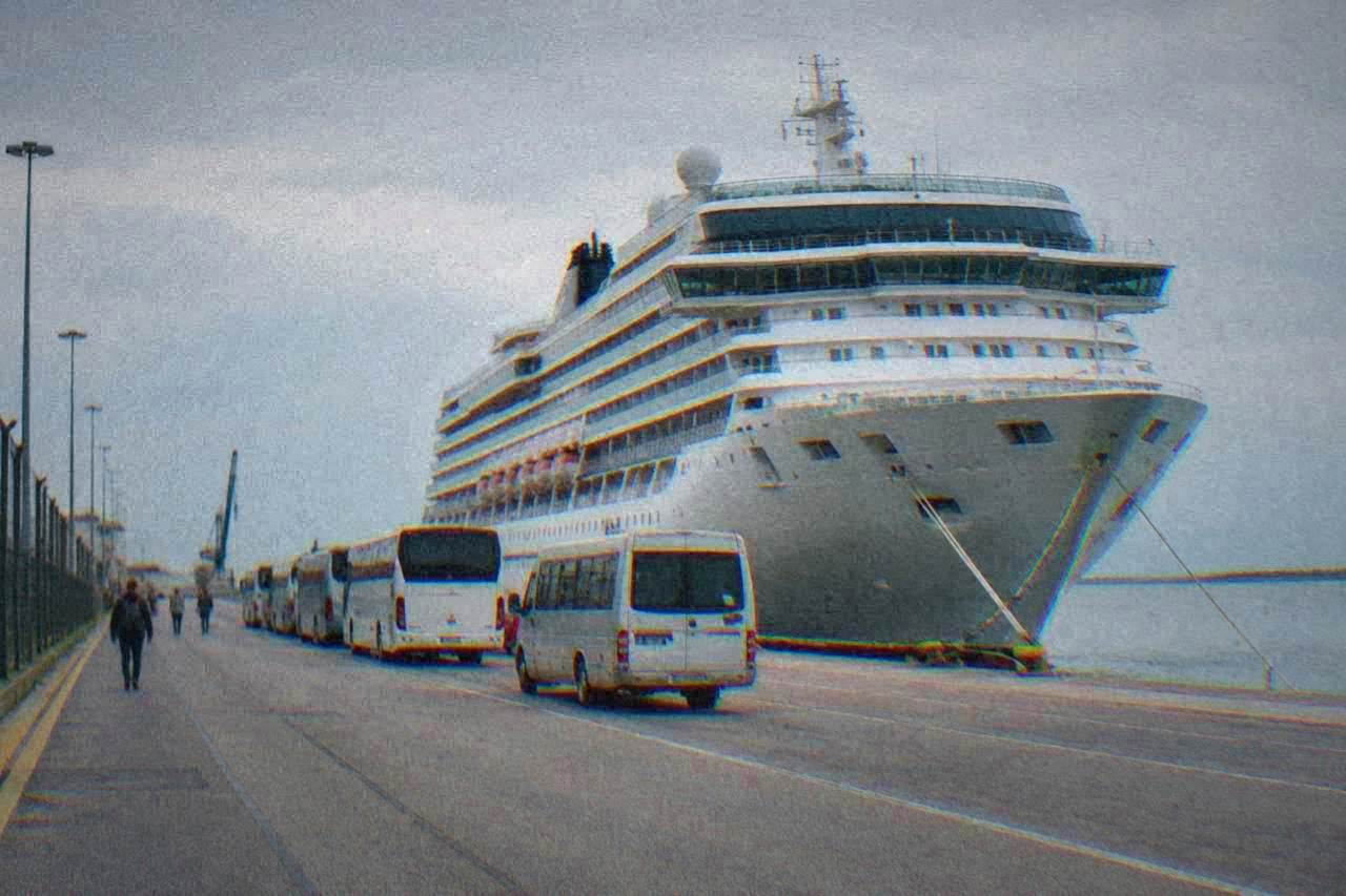 Cruise ships docked at a industrial port terminal with cranes and a shuttle bus waiting nearby