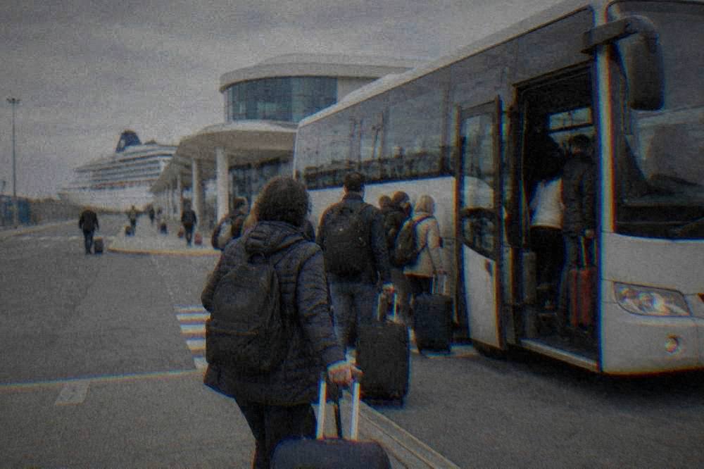 Passengers with suitcases boarding a shuttle bus outside a cruise terminal building