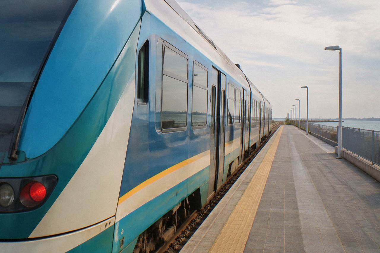 A regional train standing at a station platform beside a modest brick station building in Ravenna