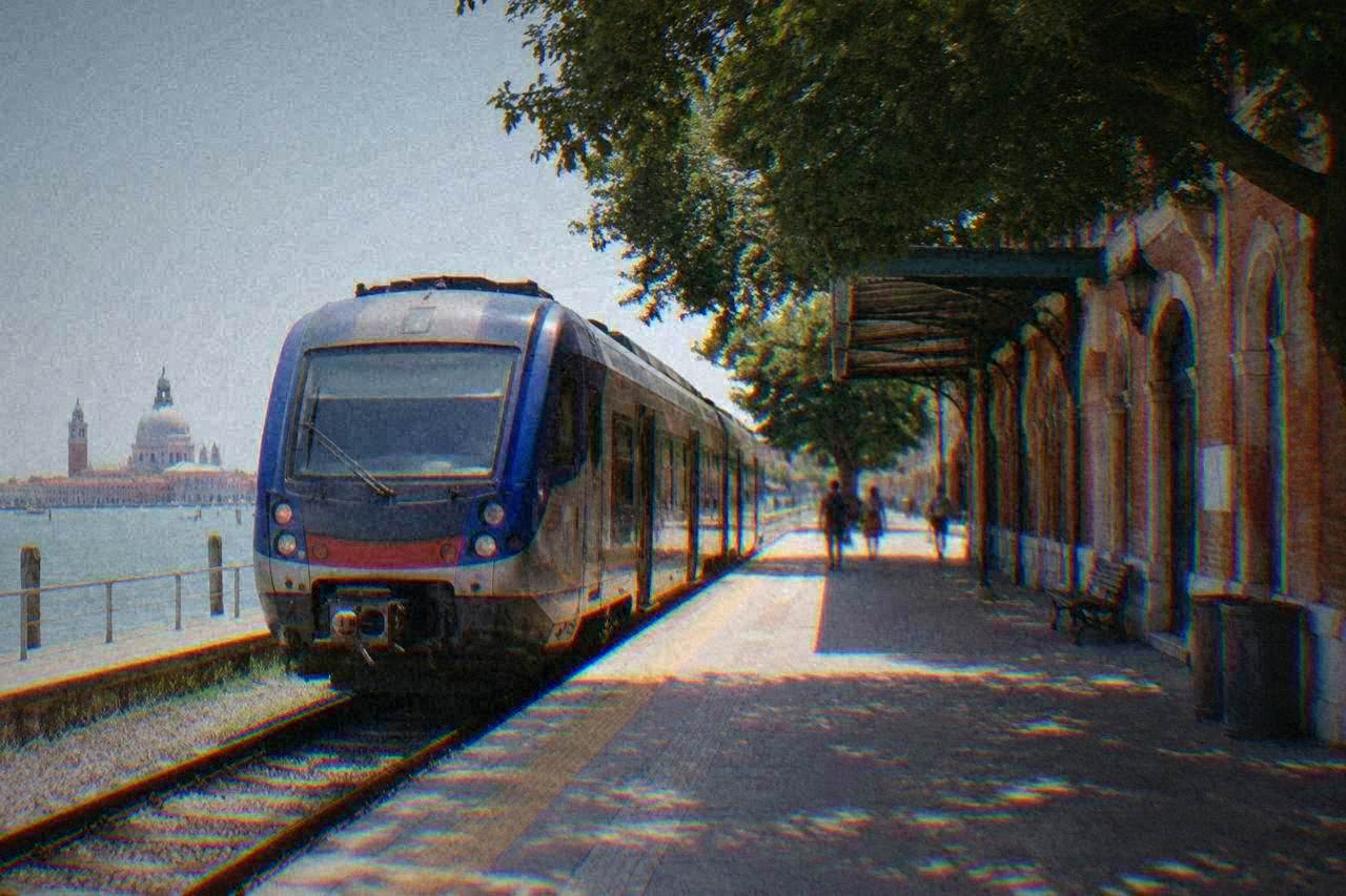 A regional train waiting at an Italian railway station platform beside a station building