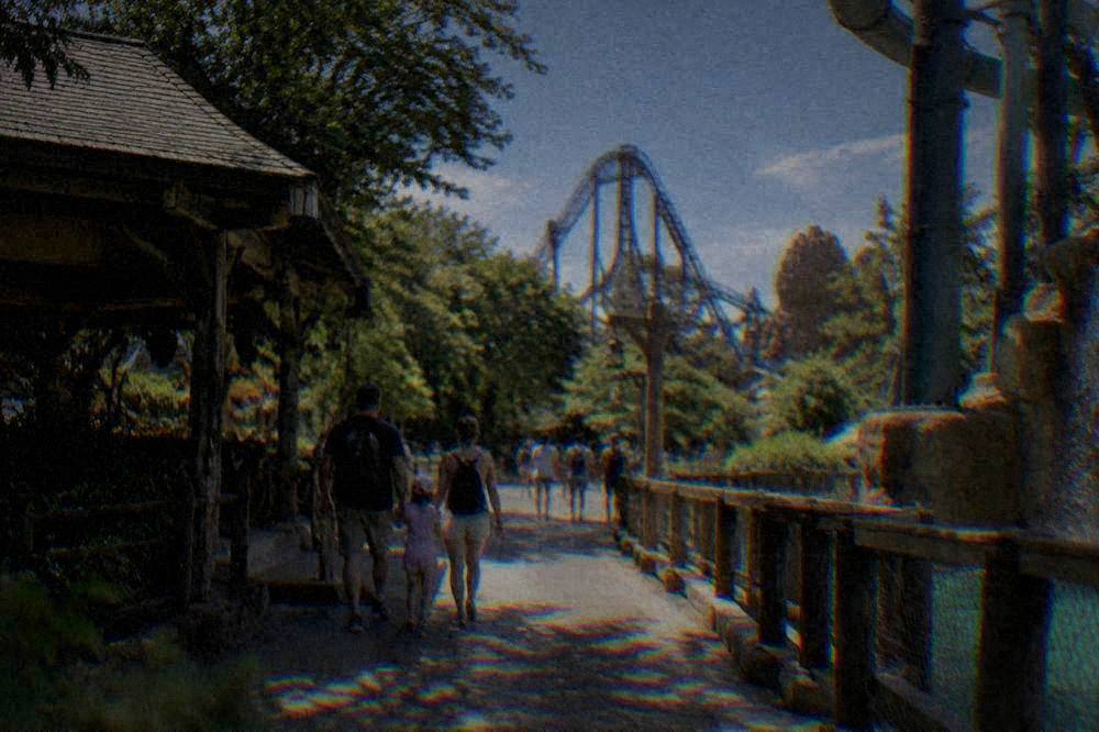 A family walking along a park path between shaded ride structures on a summer afternoon