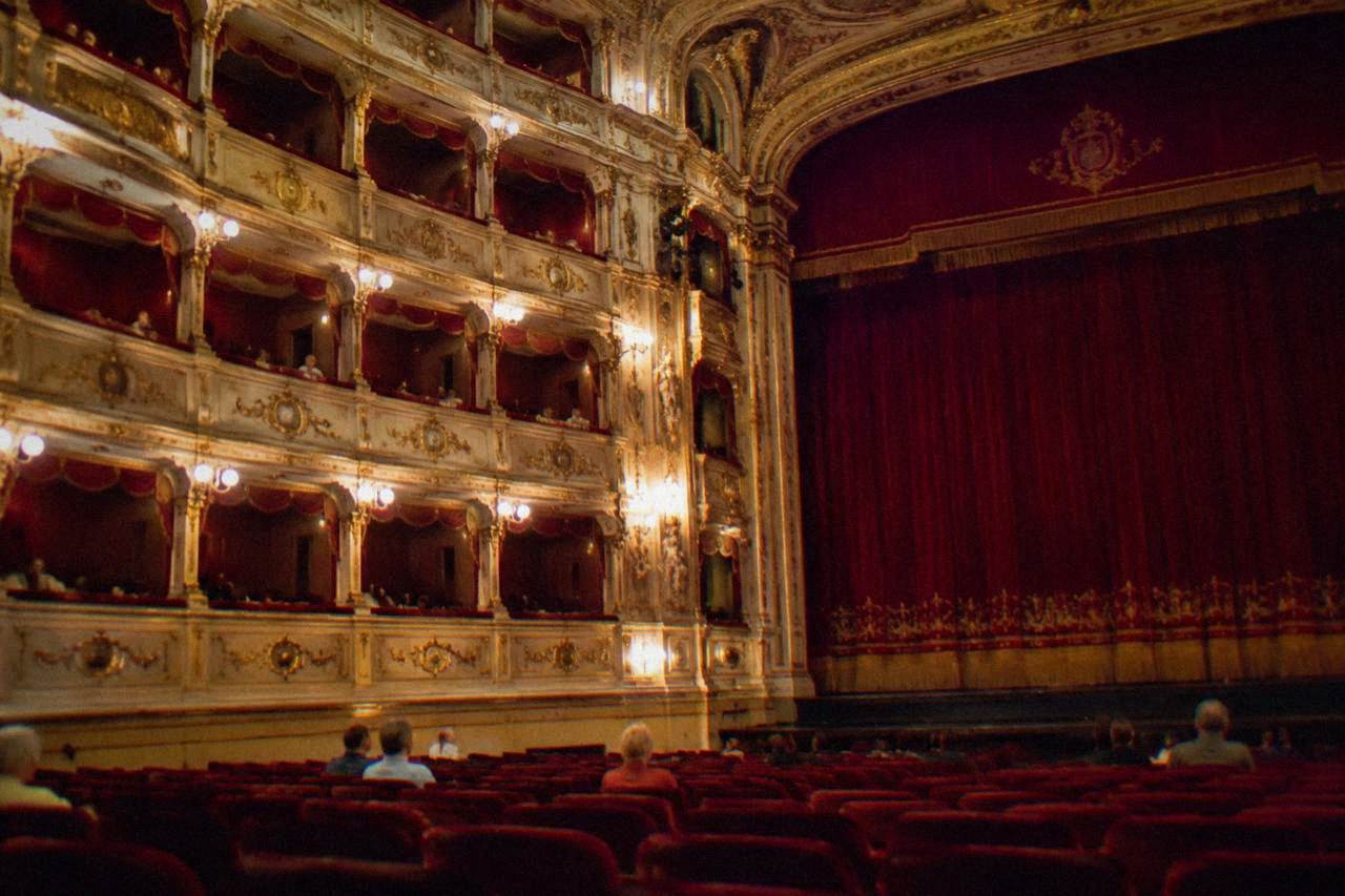An historic Italian theatre interior with rows of seats and an ornate stage lit for an evening performance
