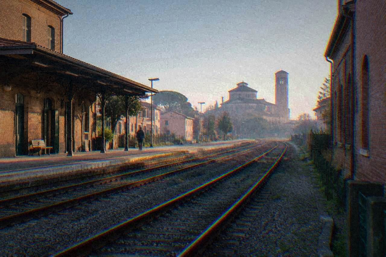 Train tracks leaving a small Italian railway station past brick buildings under a clear sky