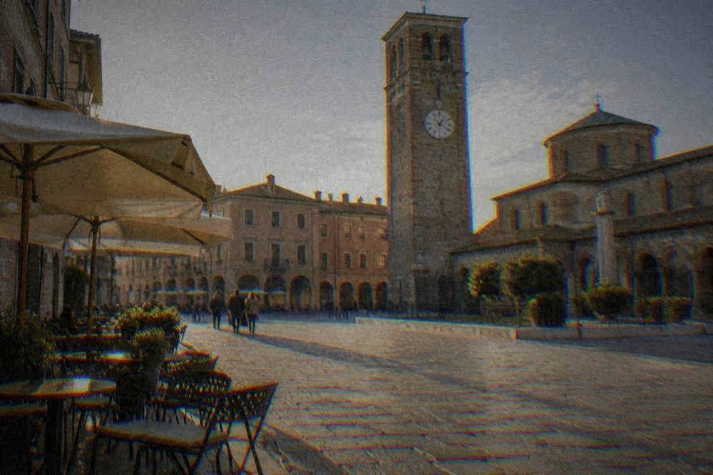 A stone-paved Italian square with a historic clock tower and café tables in morning light
