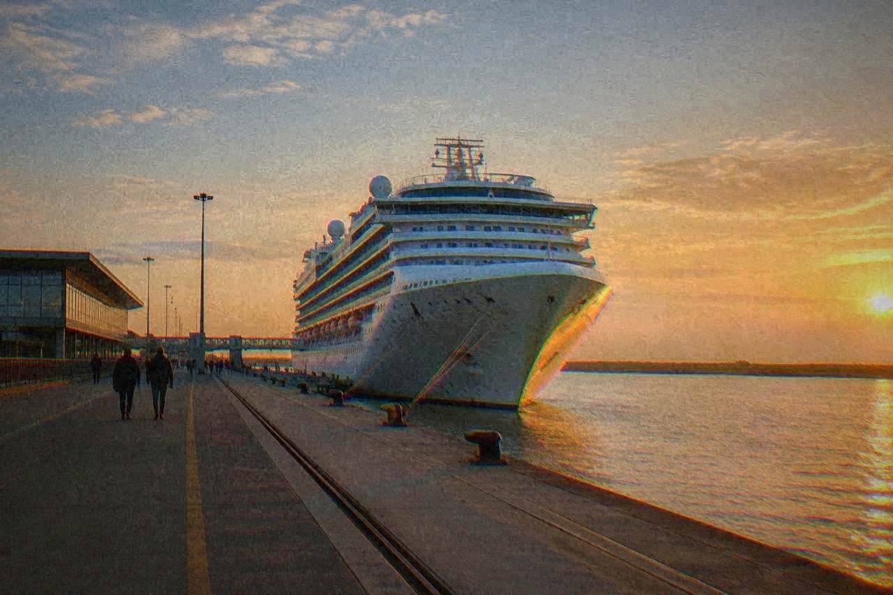 A cruise ship docked at a port terminal beside a flat Italian coastal landscape