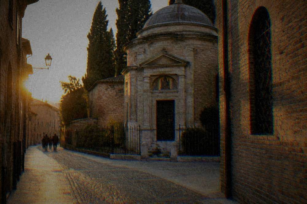 A quiet narrow street in an old Italian town with brick buildings and a small historic chapel
