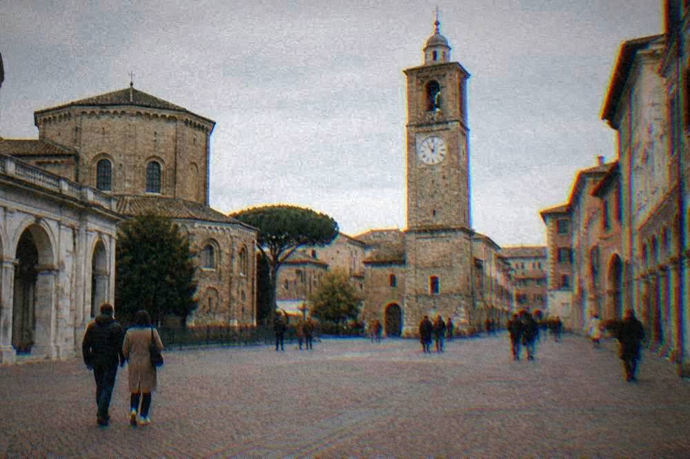 People strolling across a wide brick-paved square with a historic clock tower in Ravenna