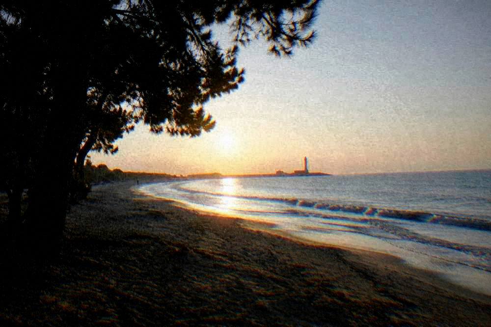 Empty beach at sunrise with gentle waves and a distant lighthouse silhouette