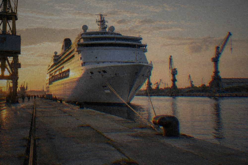 A large cruise ship moored at a industrial quay beside a canal with cranes in the background