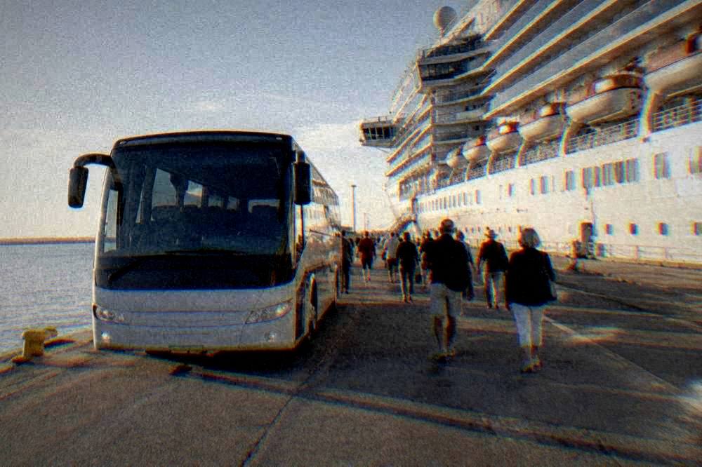 A shuttle bus parked on a quay beside a cruise ship with passengers walking toward it