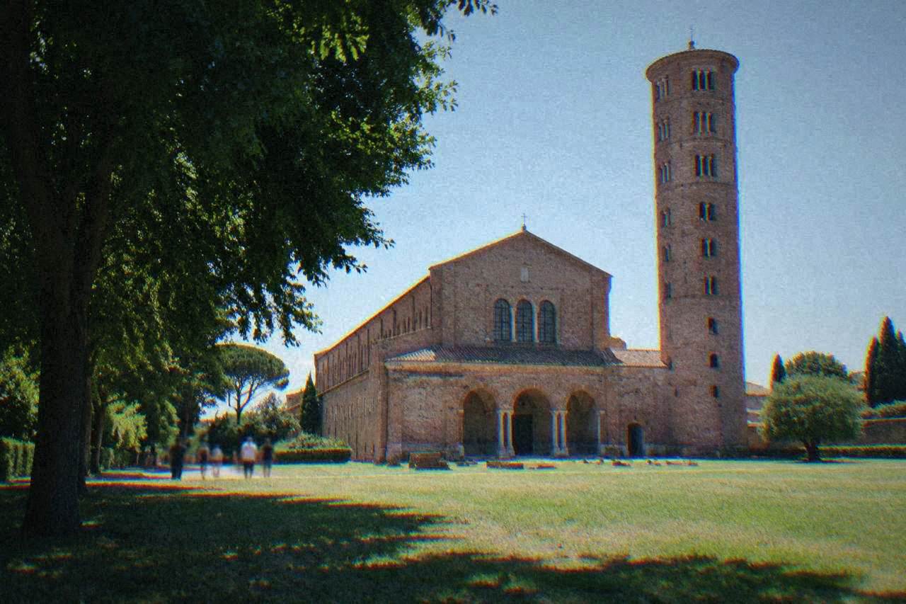 The tall brick bell tower and facade of a large basilica standing in a green field under a clear sky