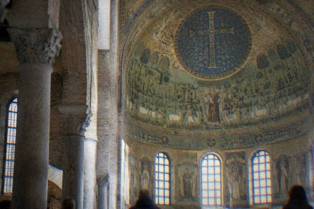 A curved apse mosaic showing a robed figure standing in a green landscape surrounded by sheep and a decorated cross