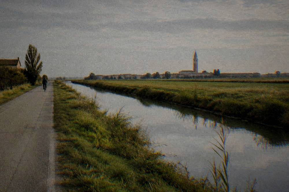 A single track road running beside a drainage canal through flat green farmland toward a distant bell tower
