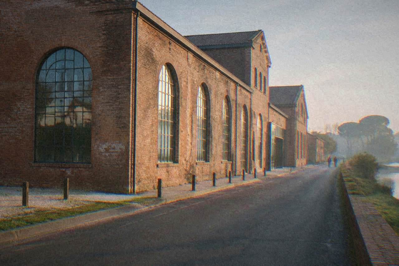 A large industrial brick building with tall arched windows standing beside a quiet road on the edge of Ravenna