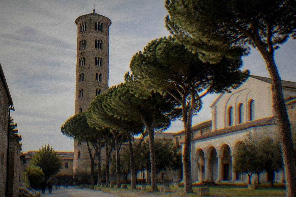 A tall brick bell tower rising above a row of stone pines beside a pale basilica facade
