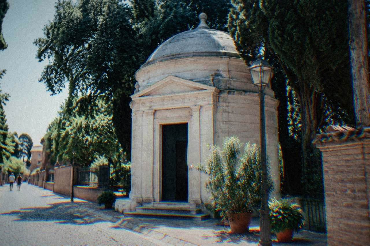 A small stone mausoleum exterior in a quiet Ravenna street, with a lamp and plants beside the doorway