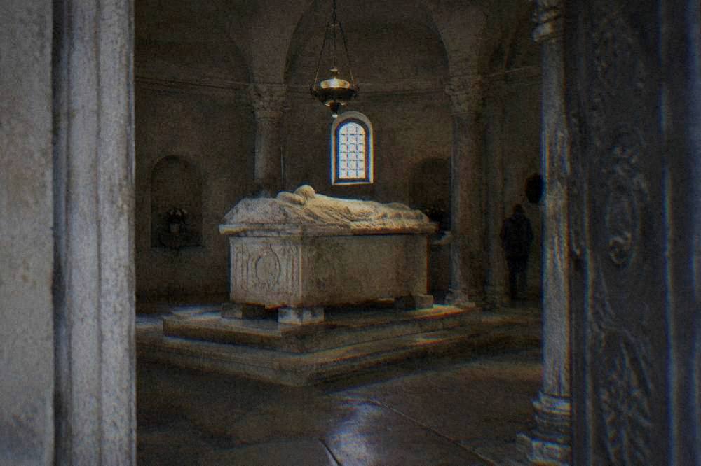 Interior of a small stone mausoleum with a raised ancient sarcophagus lit by a hanging lamp