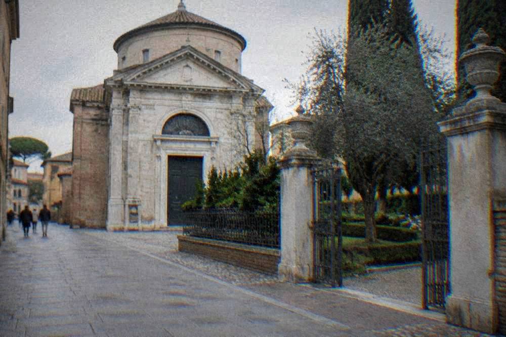 A quiet street in Ravenna with a stone church facade and a small memorial garden entrance