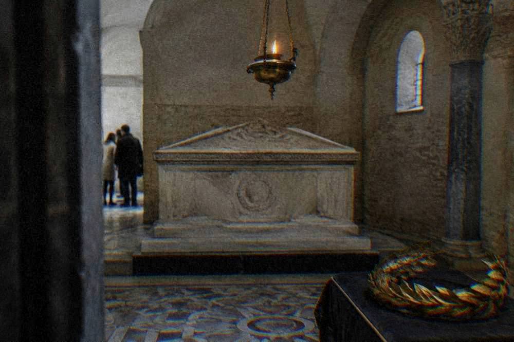 A stone tomb chamber with an oil lamp glowing above a sarcophagus and a gilded laurel wreath