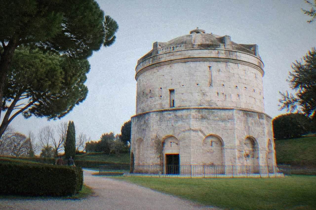 A massive round stone mausoleum of pale blocks standing in a quiet green park under a clear sky