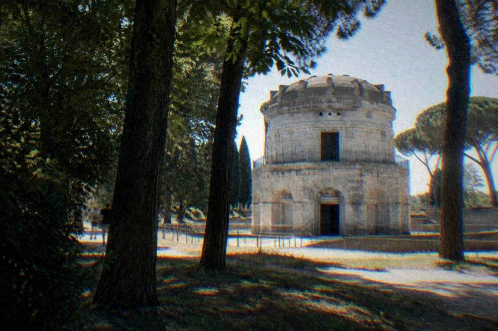 Morning light raking across the pale stone blocks of the round mausoleum seen through park trees