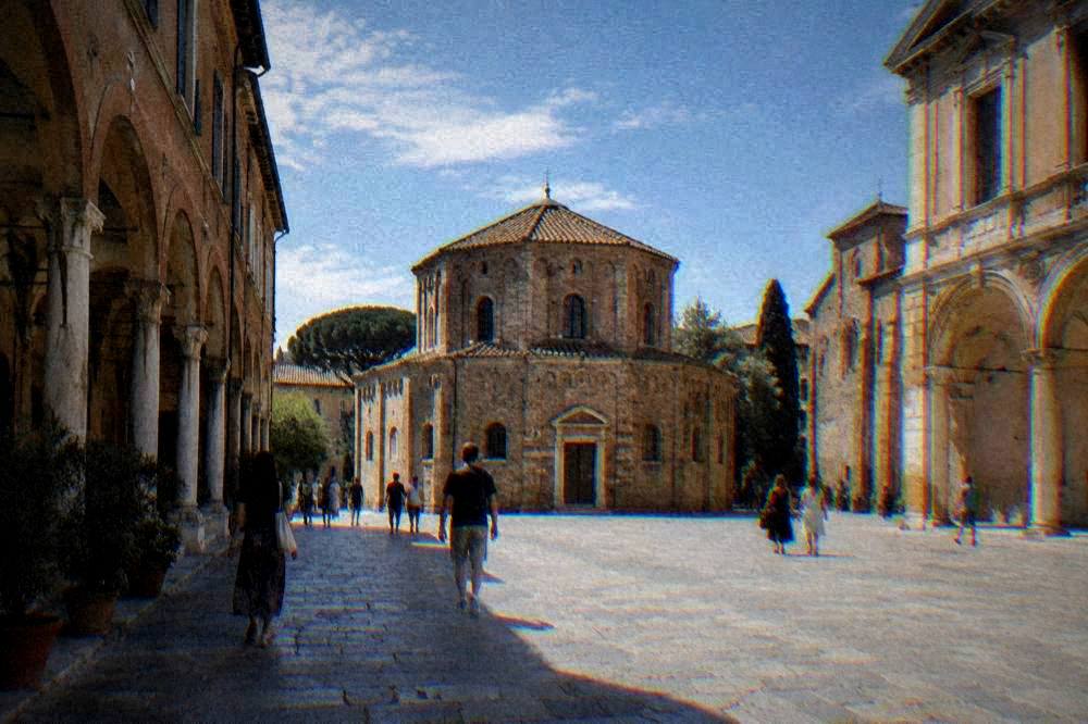 People walking across a sunlit Italian square lined with arcades and historic buildings