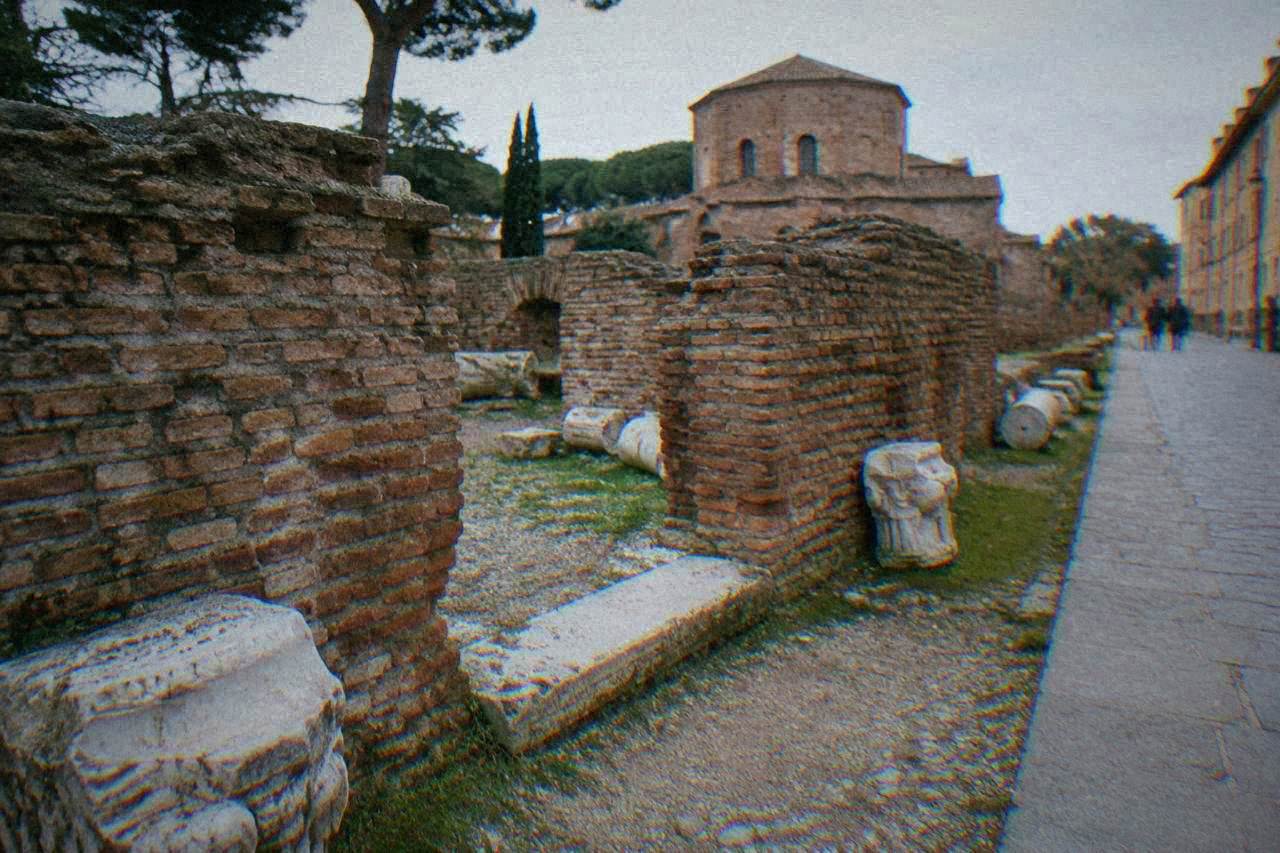 A low brick ruin with a stone threshold and scattered marble fragments beside a quiet Ravenna street