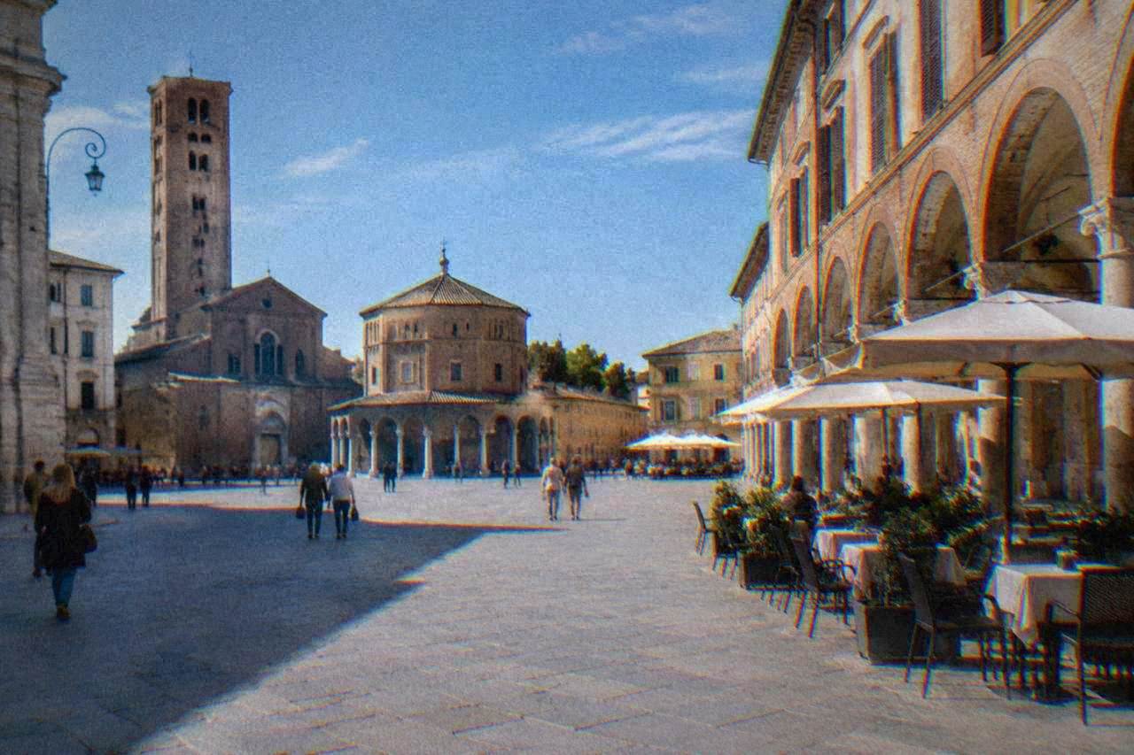 A wide paved city square lined with historic buildings and cafe tables under arcades
