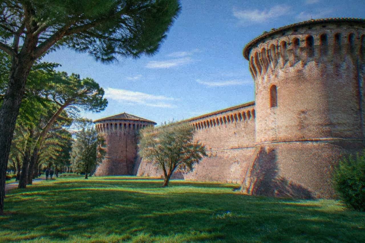 A brick fortress wall with round towers seen from a green lawn with trees in Ravenna