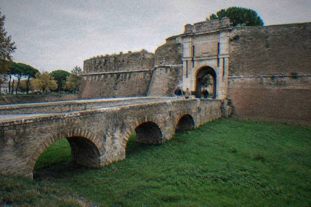 A stone bridge crossing a grassy moat toward an arched gateway in a fortress wall