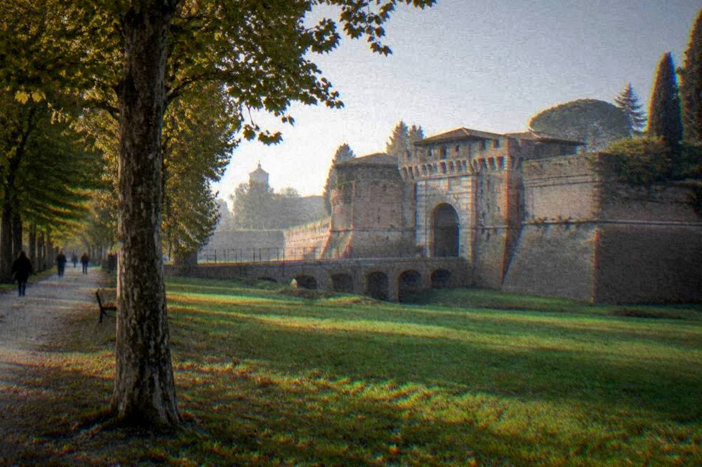 A wide lawn and tree-lined path in front of a fortress gateway in Ravenna