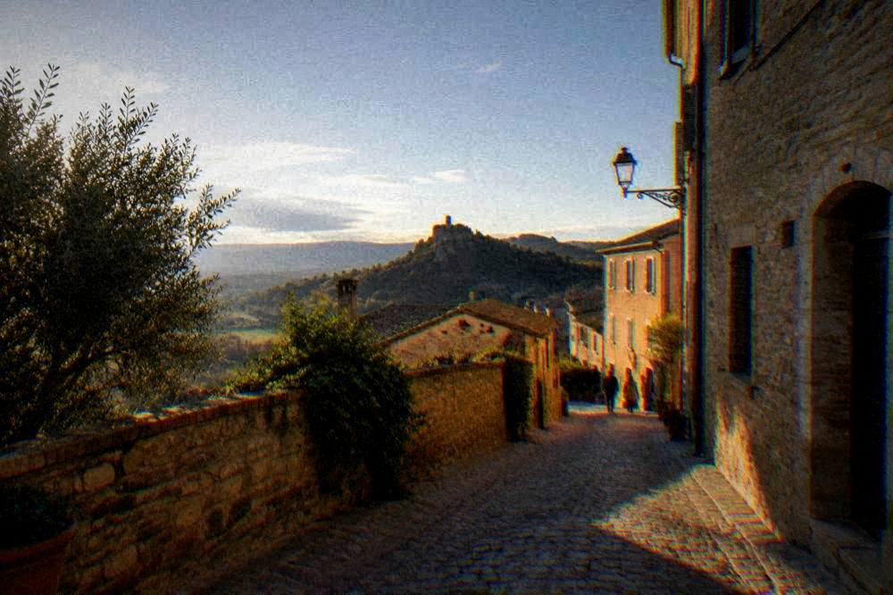 A stone clock tower on a rocky spur above the rooftops of a hill town at dusk