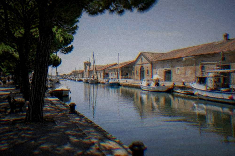 A canal in Cervia with low salt warehouses on one bank and small boats moored along the water