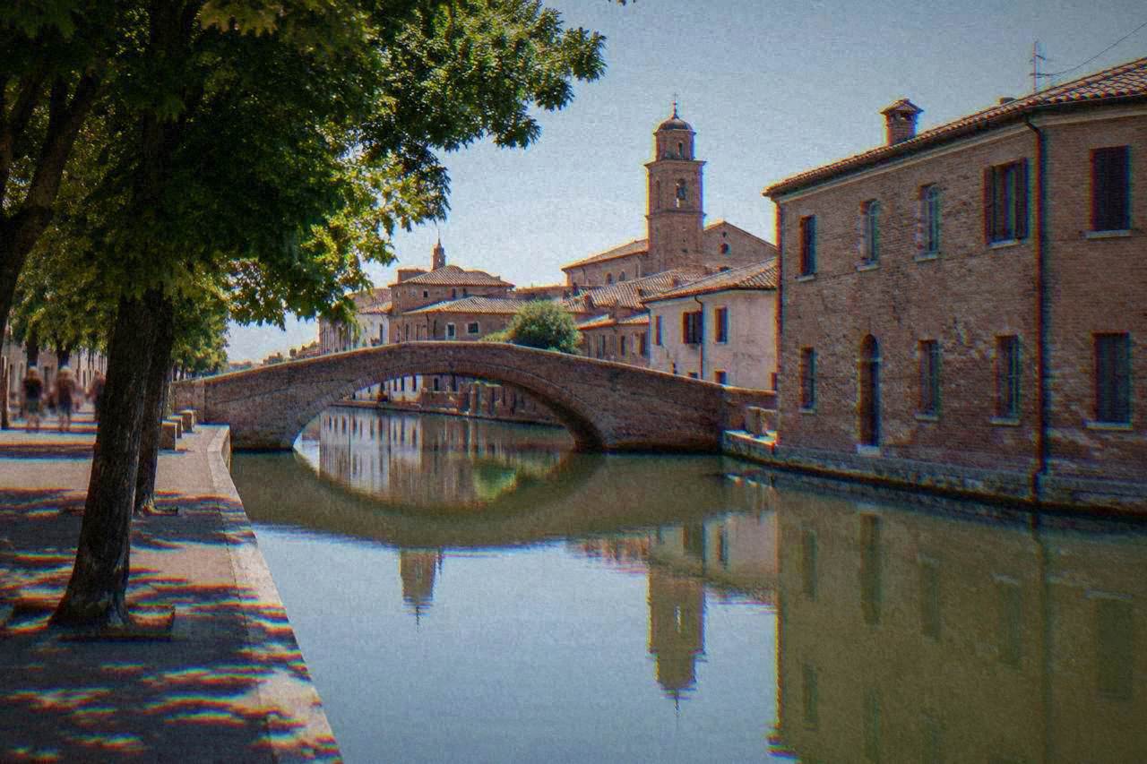 A canal in Comacchio with an arched bridge and old brick buildings reflected in still water