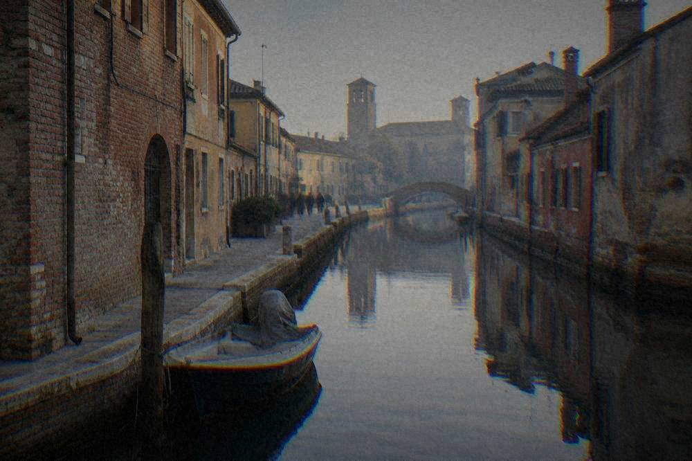 Narrow canal in Comacchio between weathered brick houses with a small boat moored at a wooden post