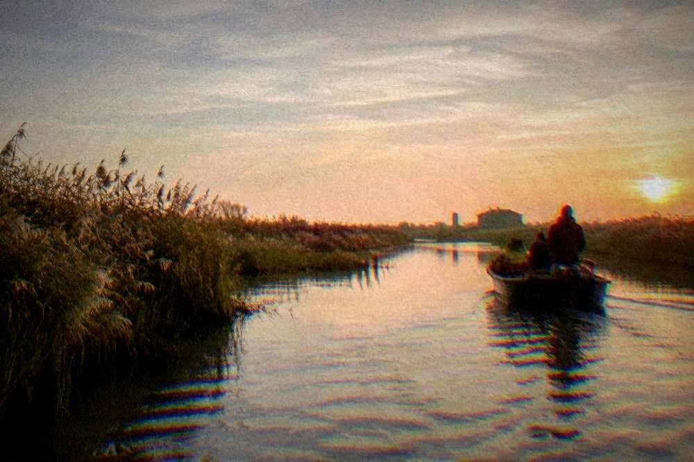 Flat-bottomed boat gliding through narrow lagoon channels past reed beds at sunrise
