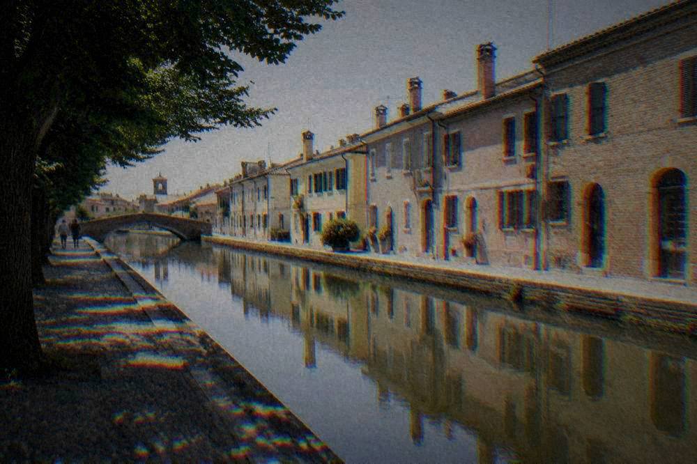 Row of brick houses reflected in a quiet Comacchio canal with a pedestrian bridge in the distance