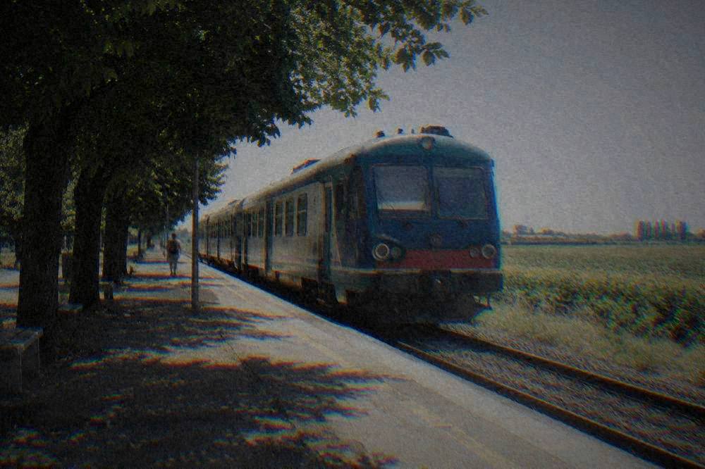 A regional train waiting at a small Italian station platform beside a flat rural landscape