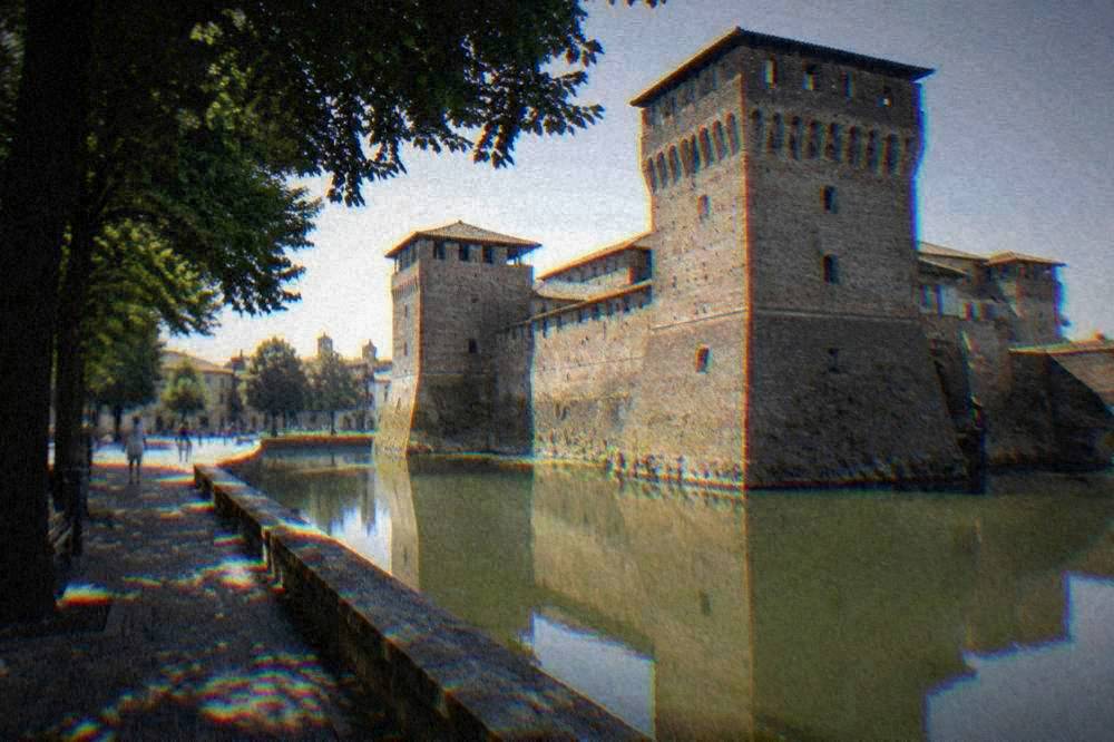 A moated brick castle with towers reflected in still water at the edge of a piazza