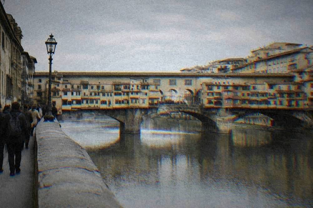 Visitors walking across an old stone bridge lined with small shopfronts over a wide river