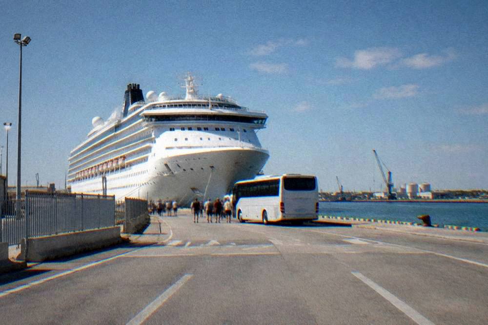 A cruise ship docked at a small industrial port with a shuttle bus waiting nearby