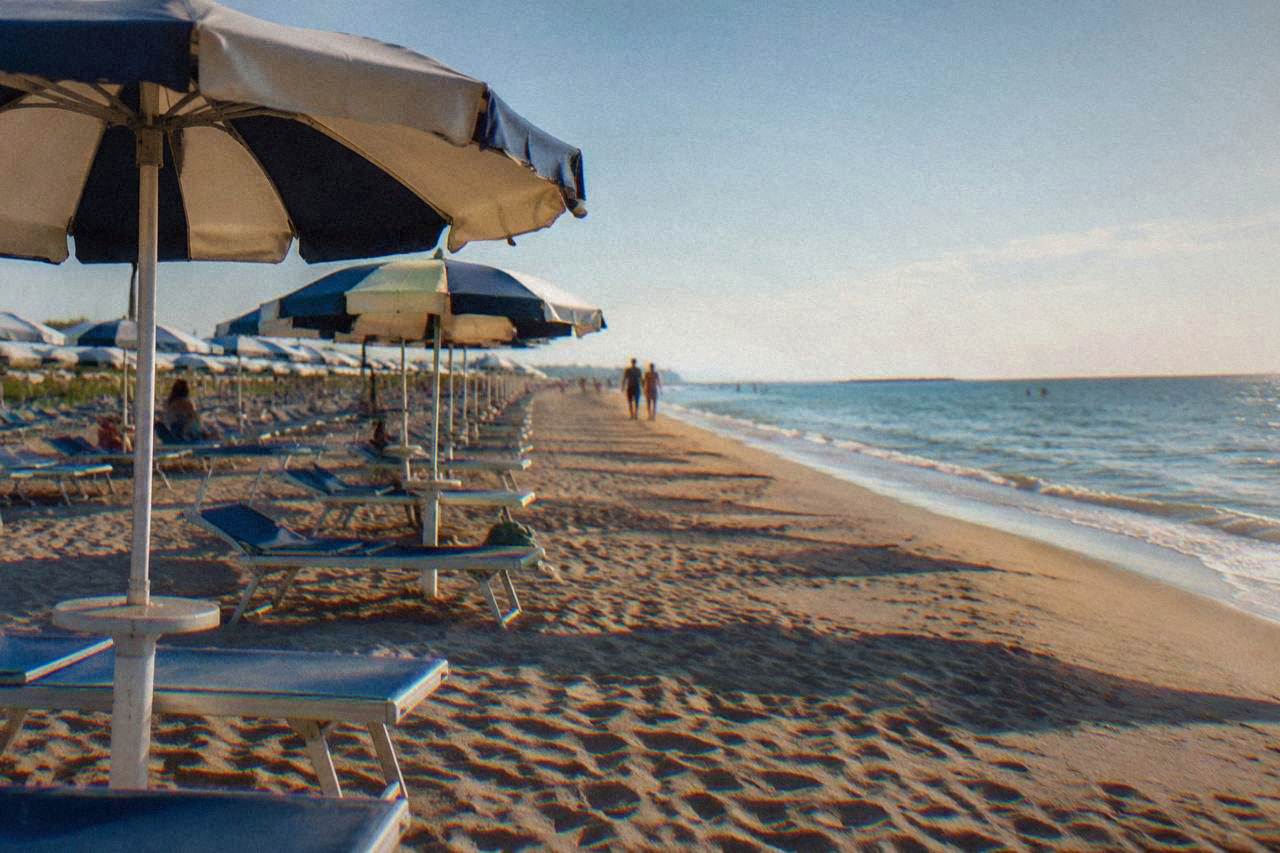 Row of umbrella-shaded sunbeds on a wide sandy beach beside a calm Adriatic sea