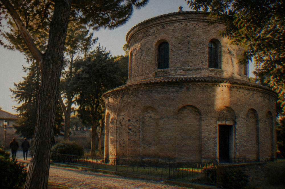 Brick exterior of a round two-storey ancient mausoleum surrounded by trees in Ravenna