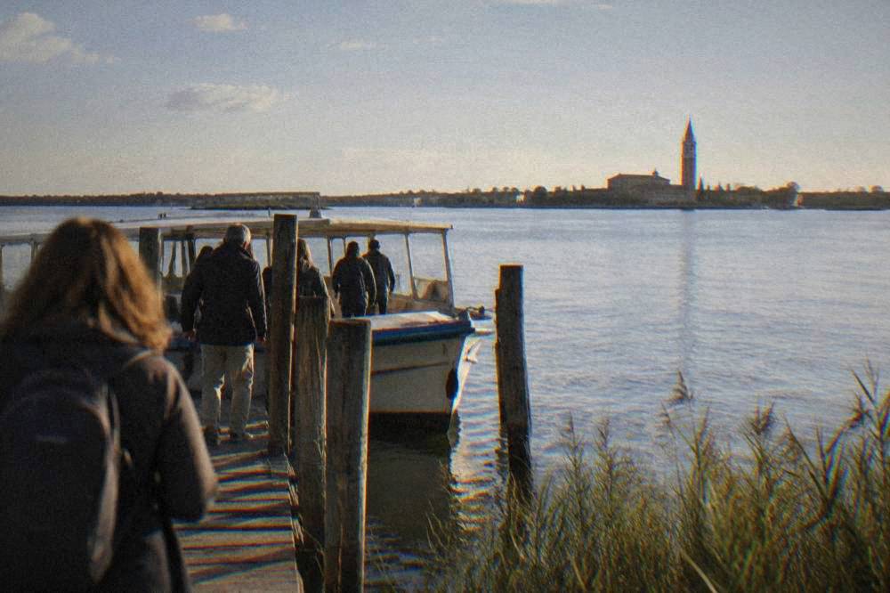 Passengers boarding a small boat on a wide lagoon with a church campanile in the distance