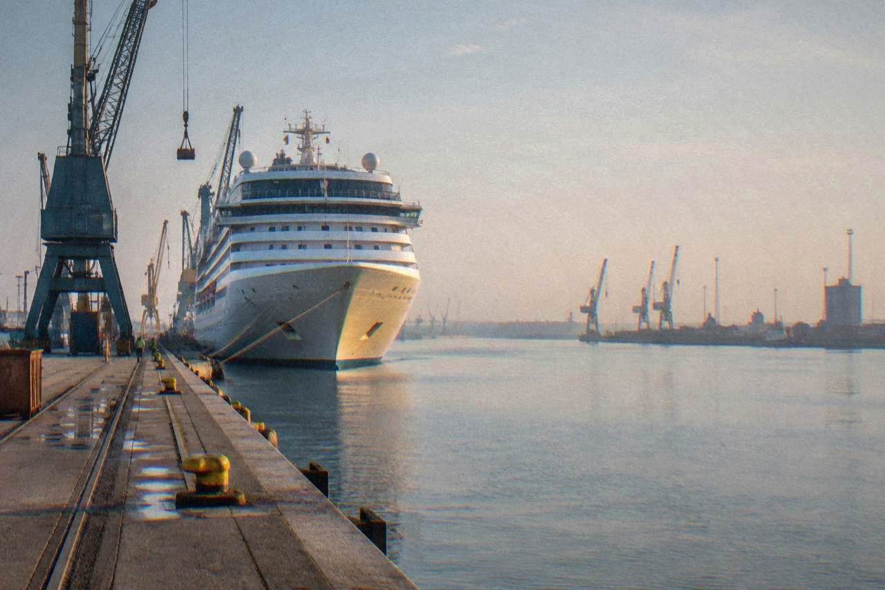 Cruise ship berthed at an industrial port terminal with cranes and a calm harbour channel