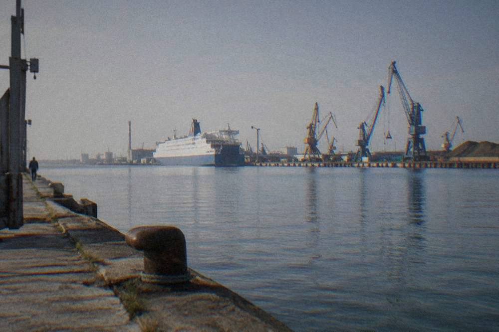 Moored ferry and port cranes seen across a wide industrial canal at midday