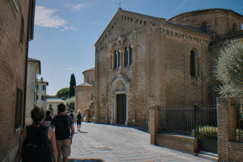 Passengers with daypacks walking past a brick basilica facade in a narrow street